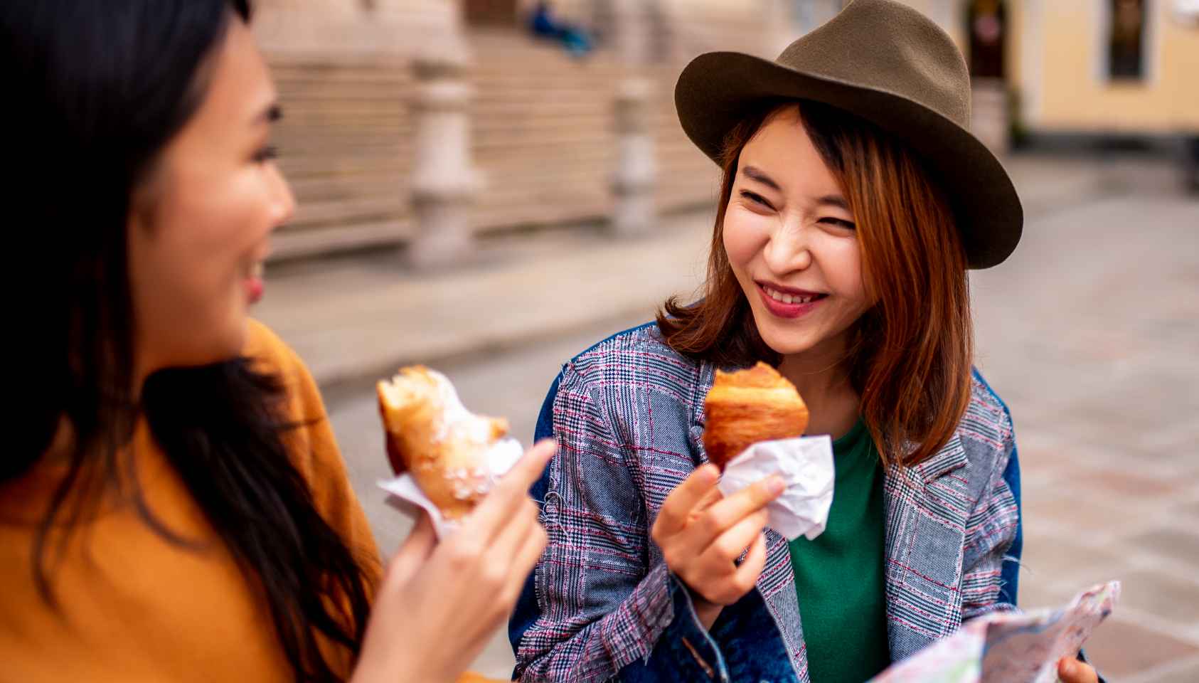 Two young women are smiling at each other while they sit outside and eat croissants